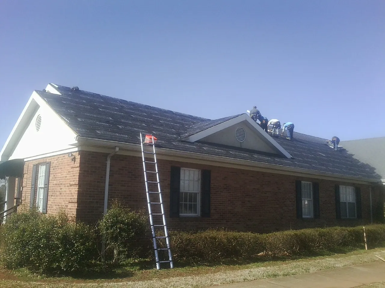Crew tearing shingles off a brick ranch roof with a ladder set — Metal Roof Repair work by our crew in Cameron