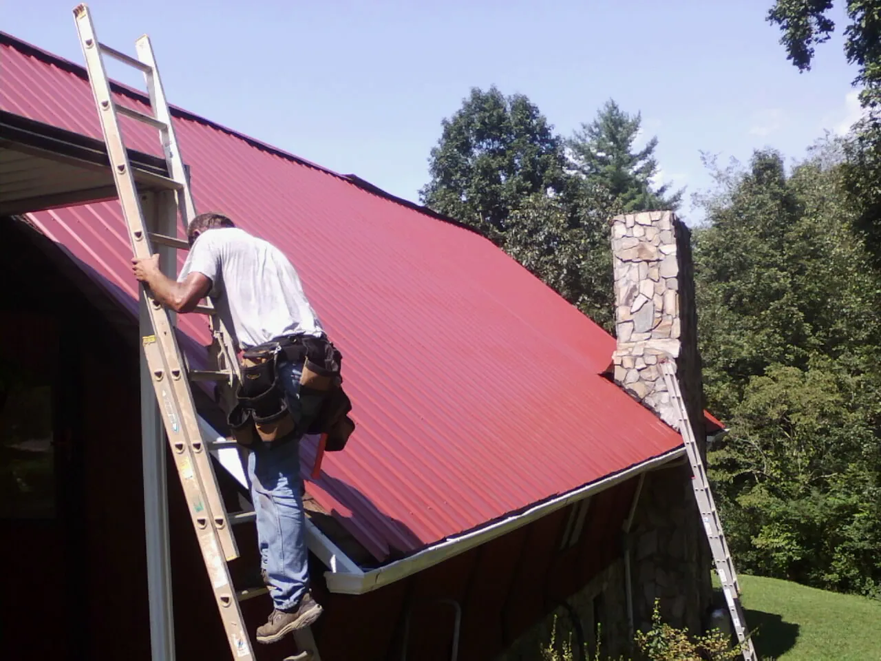 Roofer on a ladder beside a red metal roof and stone chimney — Soffit & Fascia Repair by Roofing Brothers in Cameron